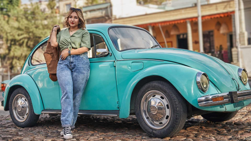 Mujer se recarga en Volkswagen Sedán clásico en tonalidad turquesa sobre calle empedrada.