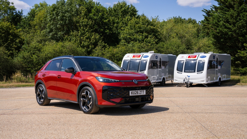 Red electric SUV parked on a sunny gravel lot beside two white travel trailers, with lush green trees behind; the scene feels calm and outdoorsy.