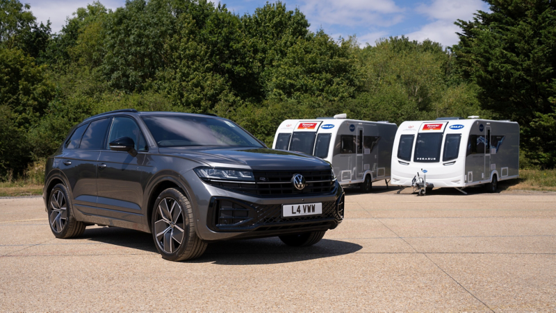 A black Volkswagen SUV is parked beside two white travel caravans on a sunlit gravel lot, with green trees behind them, creating a calm outdoor travel scene.