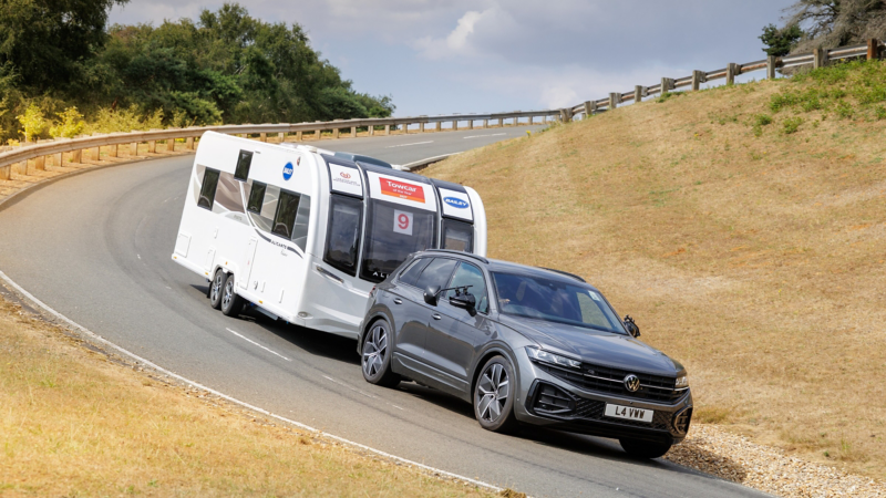 A gray Volkswagen SUV tows a white passenger trailer around a curving test road, with dry grassy slopes, trees, and wooden guardrails in daylight.