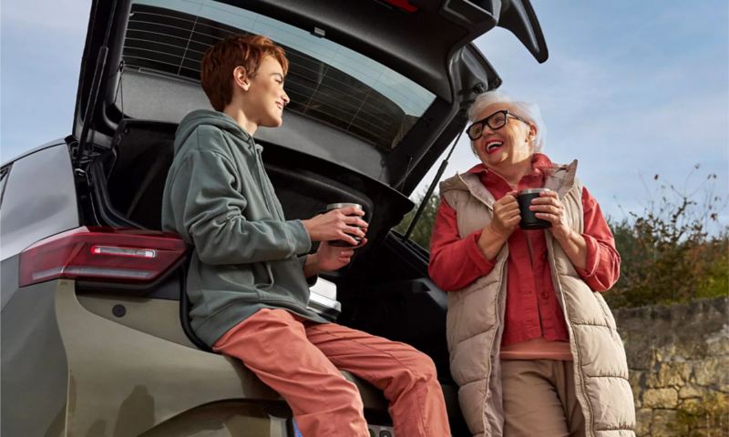 Two women enjoying a hot beverage while sitting on the tailgate of a Volkswagen vehicle.