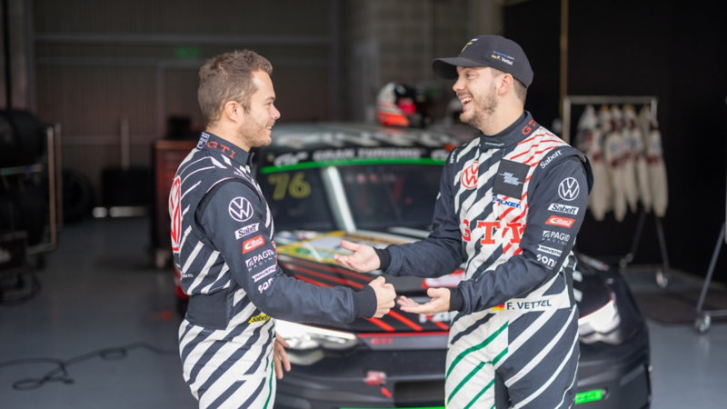Deux pilotes en combinaison Volkswagen GTI discutent dans un atelier de course devant une voiture TCR.