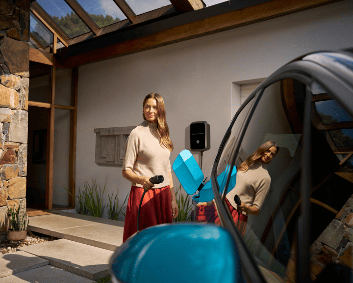 Una mujer de pie fuera de su vehículo eléctrico, sosteniendo un cable de carga con un conector en la mano.
