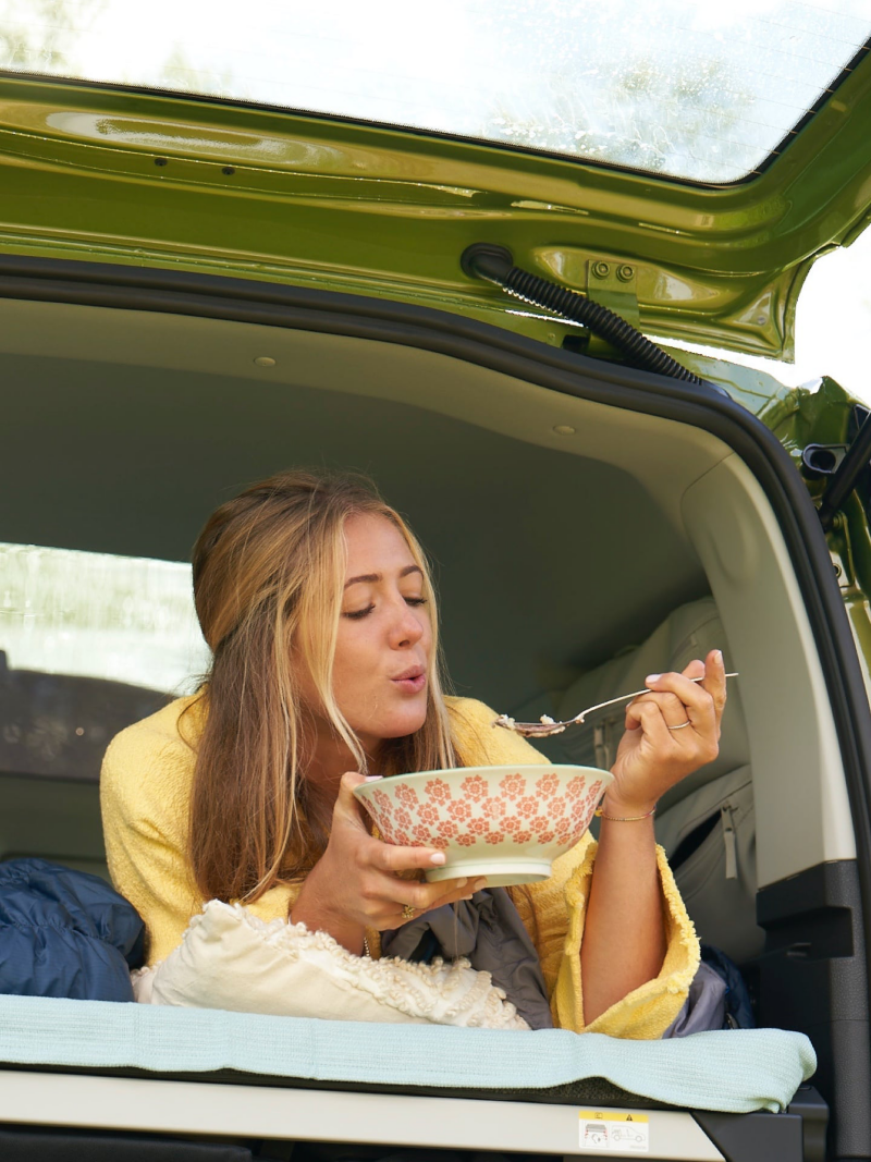 Eine Frau liegt auf einem Bett im Heck von einem VW Caddy California und isst aus einer Schüssel.