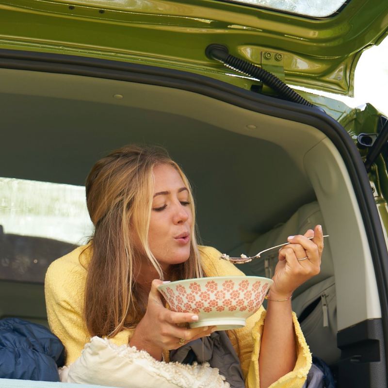 Eine Frau liegt auf einem Bett im Heck von einem VW Caddy California und isst aus einer Schüssel.