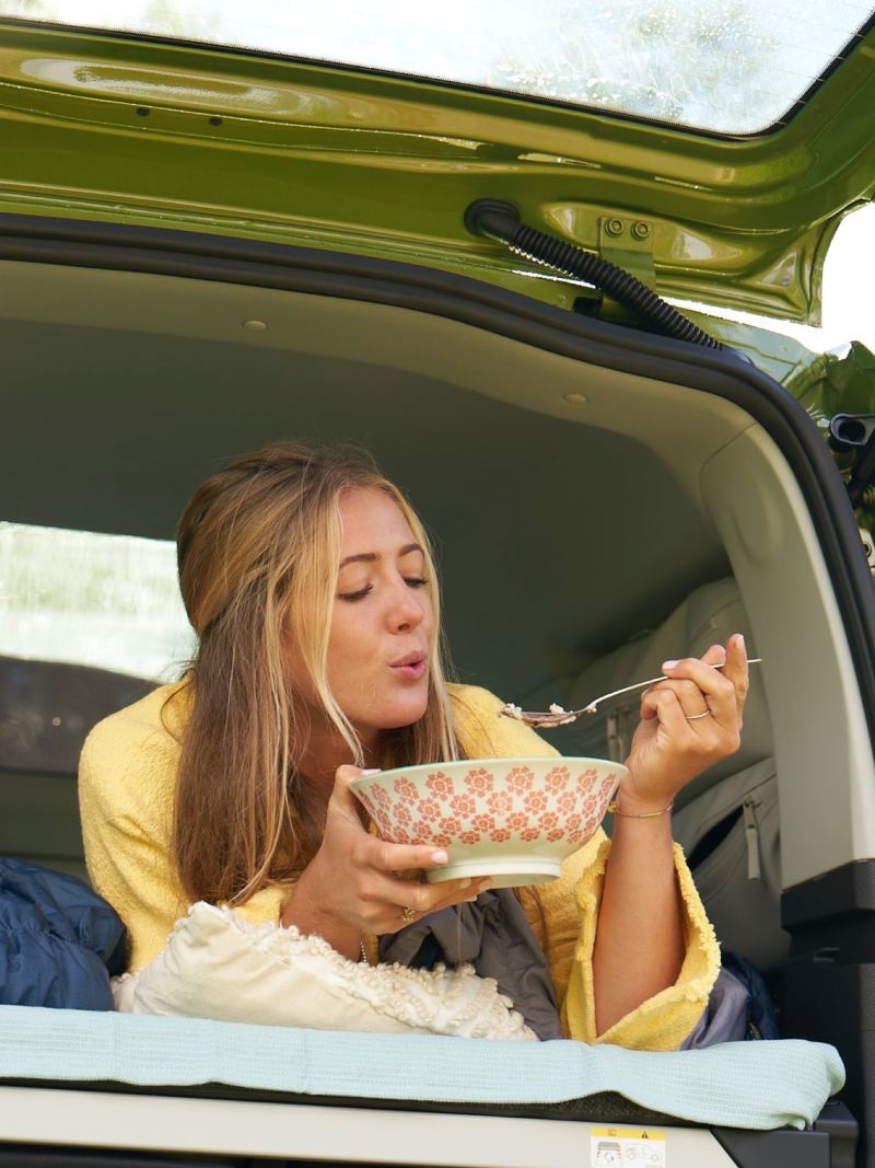 Eine Frau liegt auf einem Bett im Heck von einem VW Caddy California und isst aus einer Schüssel.