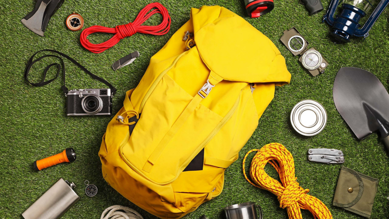 Ein Rucksack liegt am Boden umringt von verschiedensten Camping Gegenständen (Lampe, Multitool, Trinkflasche, Becher, etc.)