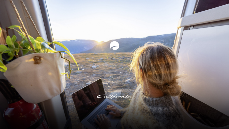 En kvinna med en laptop i knät njuter av soluppgången från sin VW Grand California husbil.