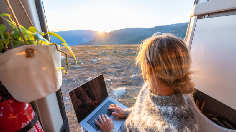 Eine Frau sitzt bei geöffneter Seitentür mit einem Laptop auf den Schoß und sieht auf die Berge.