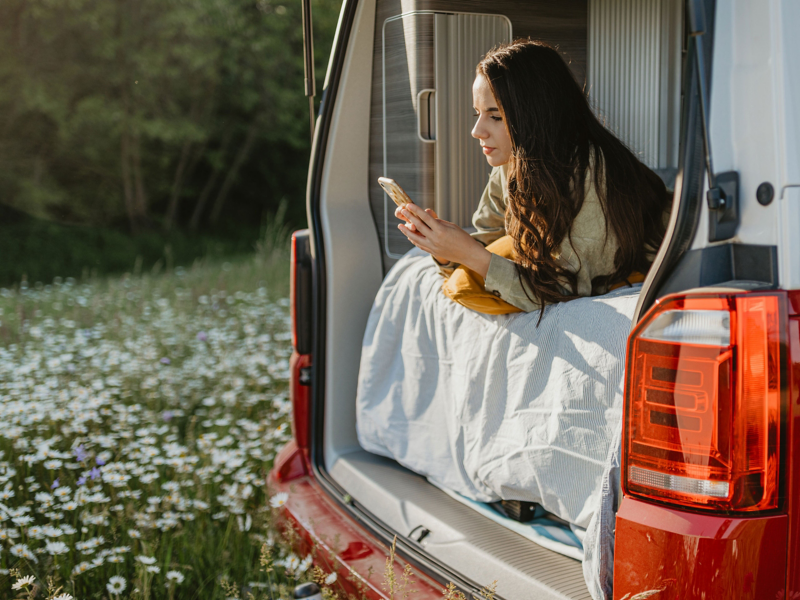 Eine Frau liegt bei geöffneter Heckklappe im Bett mit dem Smartphone in der Hand.