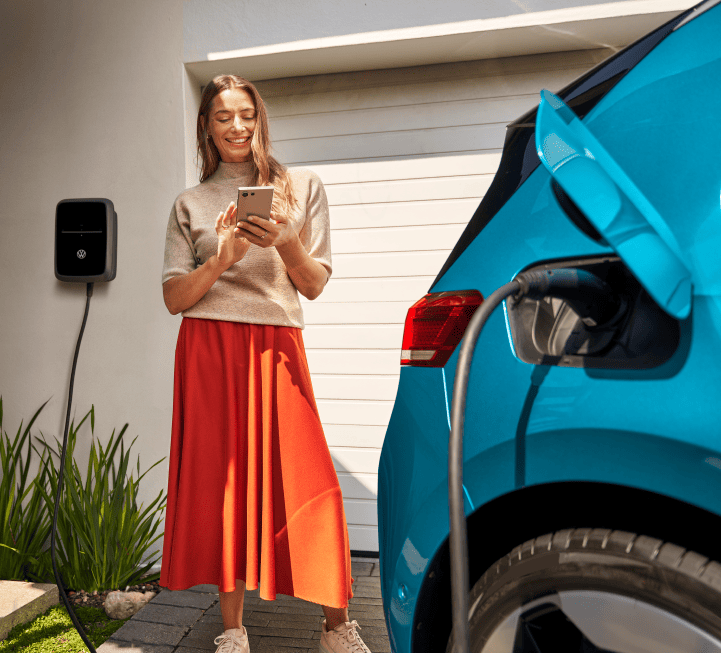 A woman standing outside her electric vehicle, holding a charging cable with a connector in her hand. She is wearing a beige sweater and a red skirt, with a charging station visible in the background.