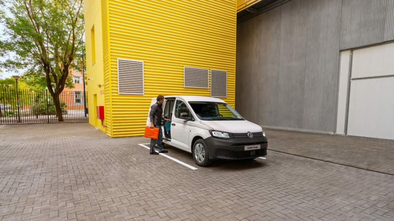 Hombre con maletín naranja sube a Volkswagen Caddy Cargo blanco estacionado frente a edificio amarillo.