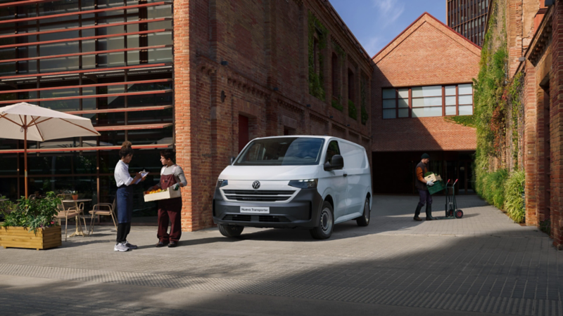 Transporter de Volkswagen blanco estacionado en patio con edificios de ladrillo. Personas conversan.