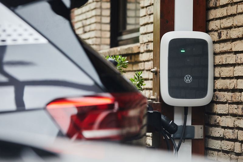 Eine Wallbox mit Volkswagen Logo hängt an einer Hauswand, im Vordergrund parkt ein Elektroauto.