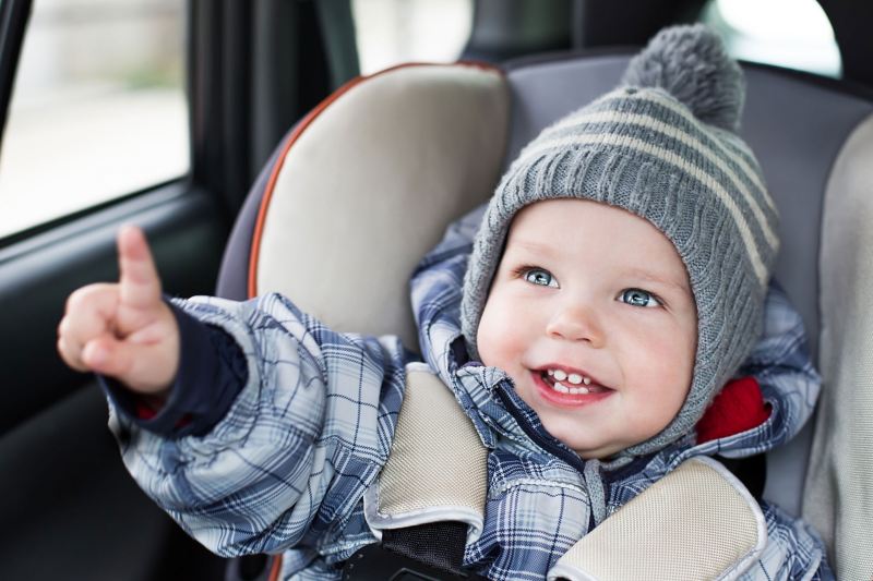 Ein gut gelauntes Kind mit Mütze sitzt in einem Kindersitz auf der Rückbank eines Fahrzeugs.