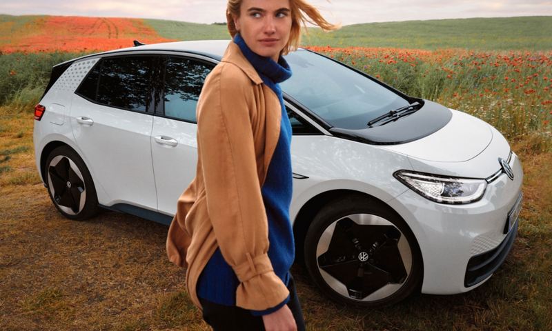 Ein weißer VW ID.3 steht auf einem Feldweg, davor schaut eine junge Frau in die Ferne. 