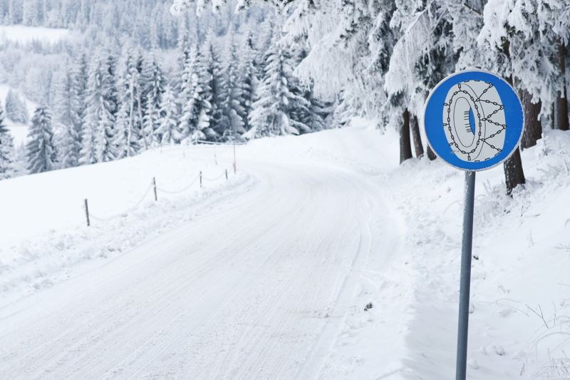 Schneebedeckte Straße und Bäume mit Verkehrsschild „Schneeketten“.