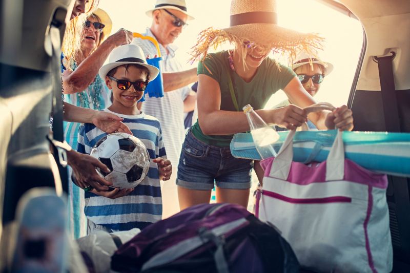 Eine große Familie mit Sonnenhüten lädt Standtaschen, einen Sonnenschirm und Strandspielzeug aus dem Kofferraum ihres Autos. 