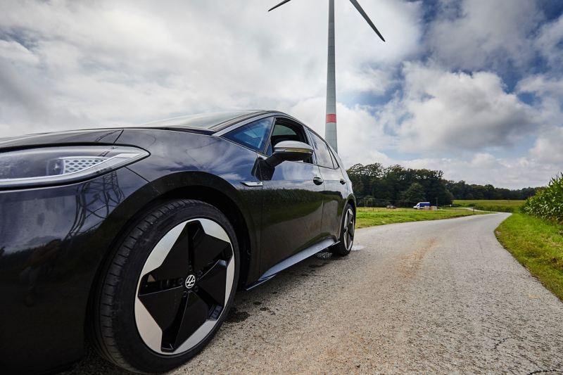Ein schwarzer VW ID.3 (Stromverbrauch kombiniert: 15,6–13,1 kWh/100 km (NEFZ); CO2-Emission kombiniert: 0 g/km; Effizienzklasse: A+++) steht auf einer Landstraße, wobei im Hintergrund ein Windrad zu sehen ist