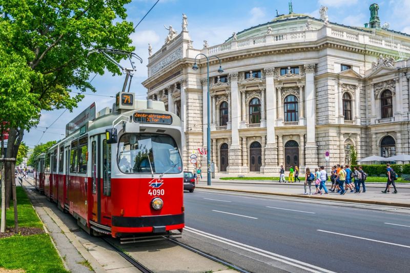 Eine historische, rote Straßenbahn fährt vor dem historischen Burgtheater in Wien die Ringstraße entlang.