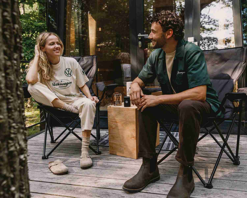 Two people sit in camping chairs; one wears a Volkswagen t-shirt, enjoying a conversation.
