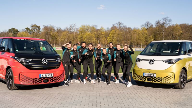 Neun Frauen der DFB Frauen Auswahl posieren in Trainingsanzügen zwischen zwei VW ID. Buzz.