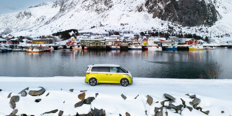 Der VW ID. Buzz fährt in einer verschneiten Landschaft vor einem See, dahinter befindet sich ein kleines Dorf.