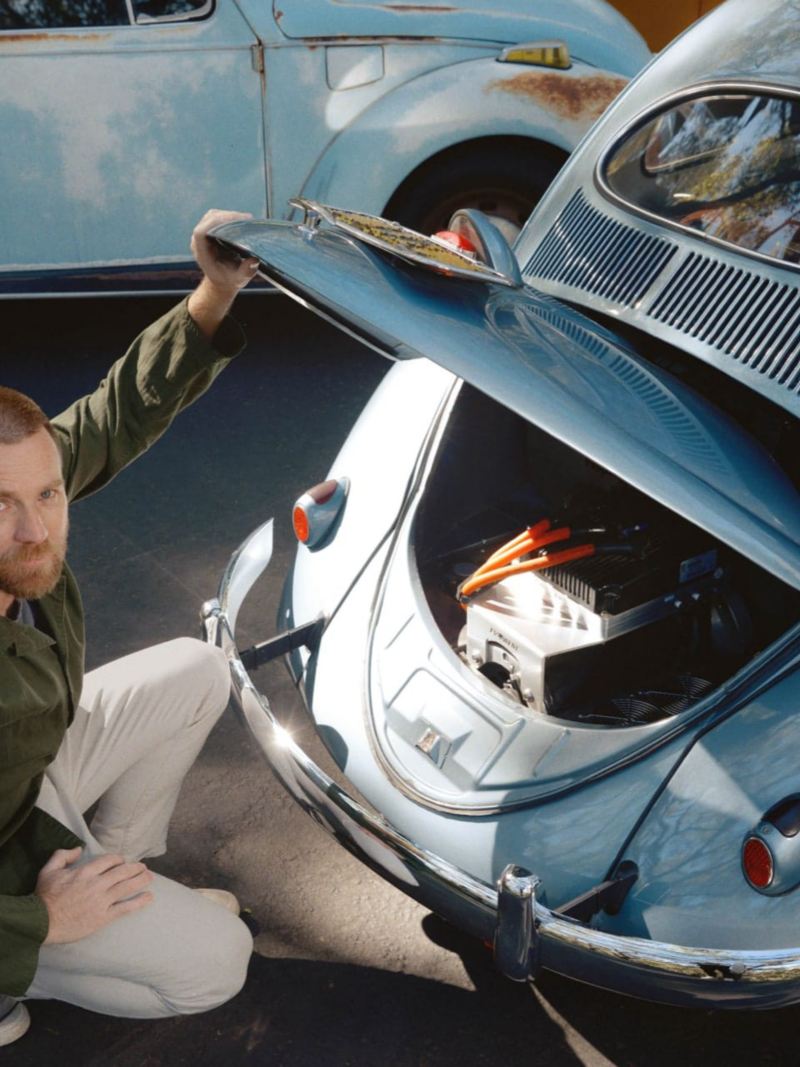 Ewan McGregor s’inginocchia davanti a un Maggiolino blu con il cofano motore aperto