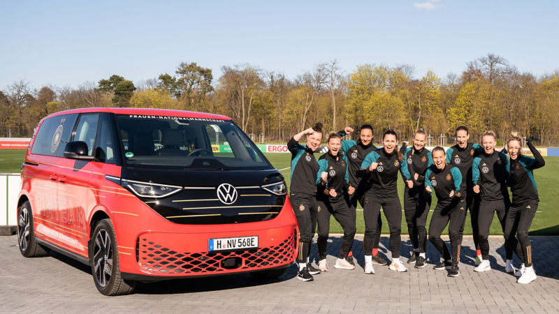 Anfeuernde DFB Frauen neben einem VW ID. Buzz mit Sonderfolierung.