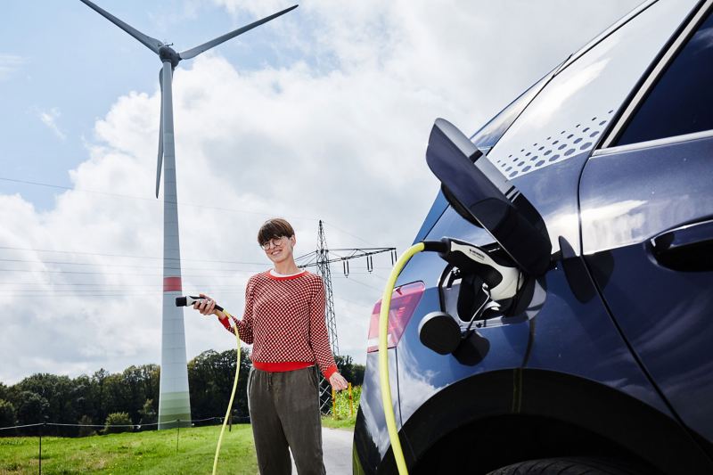 Eine junge Frau steckt das Ladekabel eines VW ID.3 (Stromverbrauch kombiniert: 15,6–13,1 kWh/100 km (NEFZ); CO2-Emission kombiniert: 0 g/km; Effizienzklasse: A+++) symbolisch in ein Windrad. 