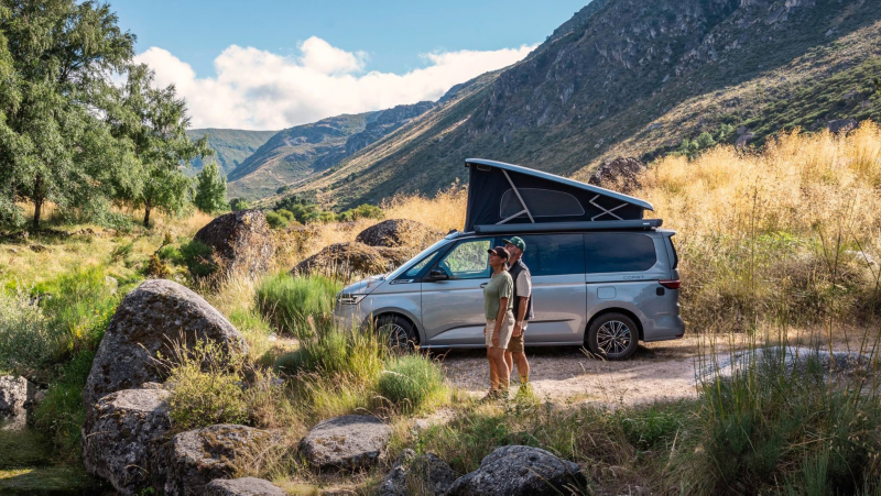 Silberner Volkswagen Camper mit Aufstelldach steht neben einem klaren Bergbach, zwei Personen betrachten die Landschaft in einer hügeligen Naturkulisse.