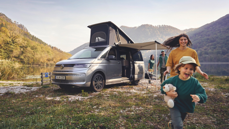 Camping-car Volkswagen argenté avec toit relevable stationné au bord d’un lac en paysage montagneux, famille se déplaçant devant le véhicule.