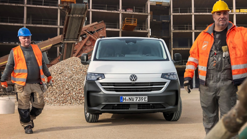 Volkswagen Transporter steht auf einer Baustelle vor einem Rohbau, Bauarbeiter mit Schutzhelmen und Warnwesten arbeiten im Vordergrund.