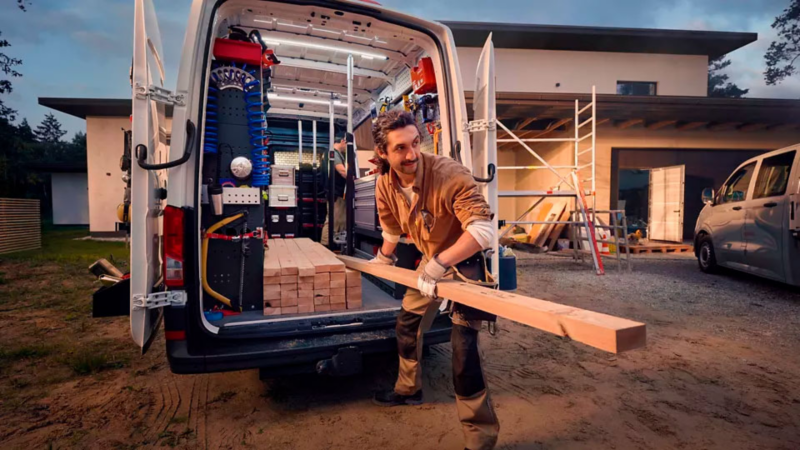 Un artisan retire des poutres en bois d'un Volkswagen Transporter ouvert, garé devant un bâtiment moderne entouré d'échafaudages.