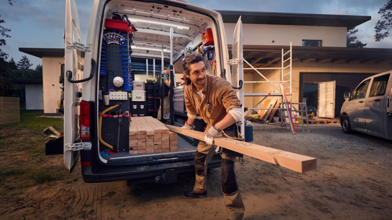 Handwerker entnimmt Holzbalken aus einem offenen Volkswagen Transporter vor einem modernen Gebäude mit Baustellengerüst.