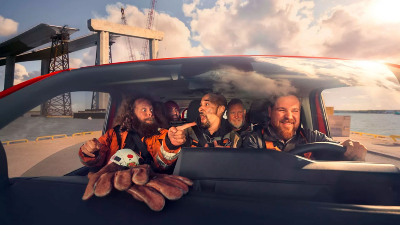 Plusieurs ouvriers assis à l’intérieur d’un utilitaire Volkswagen rouge, rient et discutent pendant le trajet, installation industrielle en arrière-plan.