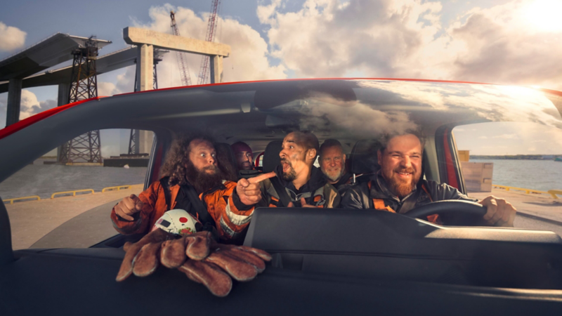Mehrere Arbeiter sitzen im Innenraum eines roten Volkswagen Transporters, lachen und unterhalten sich während der Fahrt, Industrieanlage im Hintergrund.