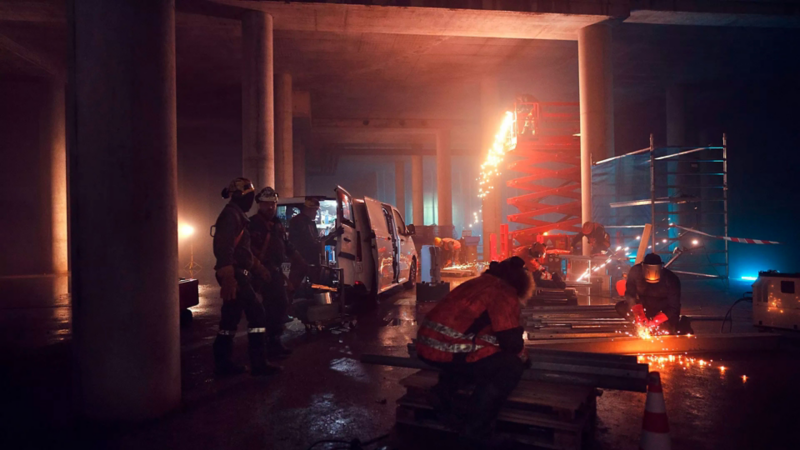 Plusieurs ouvriers travaillent de nuit sur un chantier sous un pont. Des étincelles lors de la découpe de métal illuminent la scène, tandis qu’un Volkswagen Véhicules Utilitaires Transporter est garé à côté avec la porte coulissante ouverte.