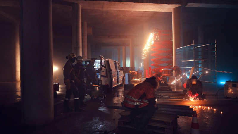 Plusieurs ouvriers travaillent de nuit sur un chantier sous un pont. Des étincelles lors de la découpe de métal illuminent la scène, tandis qu’un Volkswagen Véhicules Utilitaires Transporter est garé à côté avec la porte coulissante ouverte.