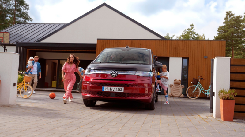 Roter Volkswagen Van steht in der Einfahrt eines modernen Hauses, Familie mit Kindern und Fahrrädern im Vordergrund.