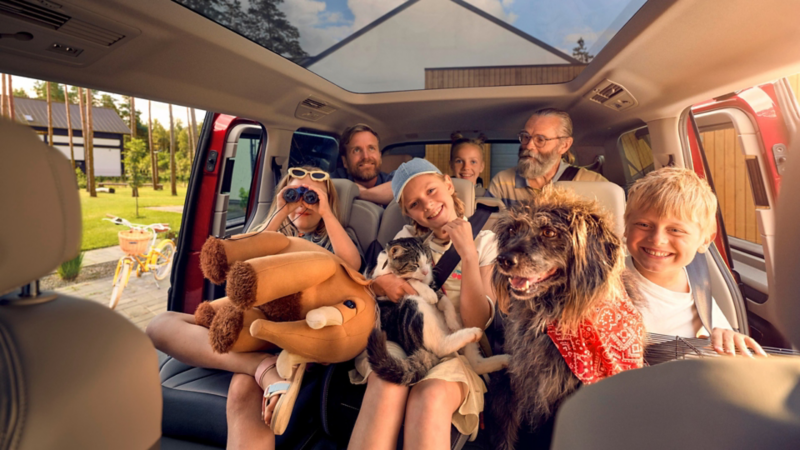Famille avec enfants, chien et chat assise à l’intérieur d’un van Volkswagen rouge avec toit panoramique.
