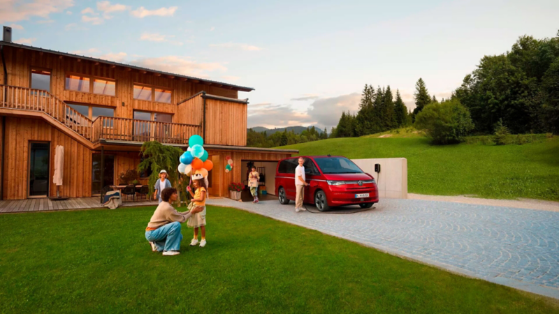 Une famille se tient devant une maison moderne en bois à côté d’un VW Multivan rouge, tandis qu’un enfant tient des ballons et que le père se trouve près du véhicule.