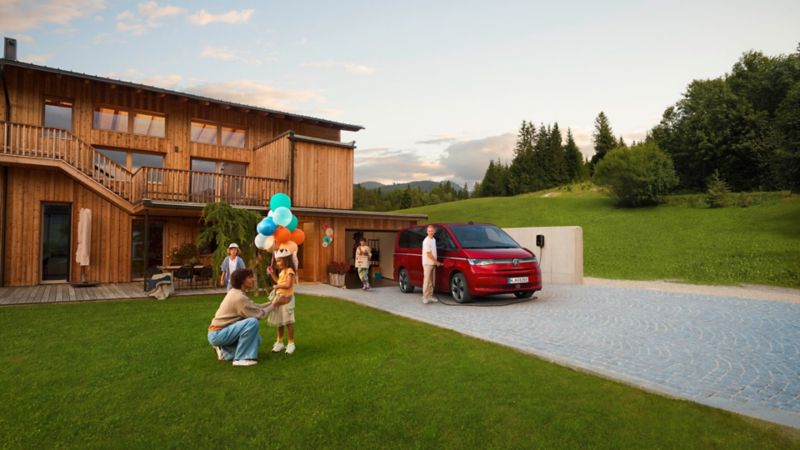 Eine Familie steht vor einem modernen Holzhaus neben einem roten VW Multivan, während ein Kind Luftballons hält und der Vater am Fahrzeug steht.