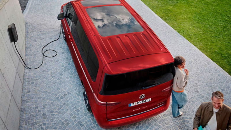 Vue aérienne d’un VW Multivan rouge en charge devant une maison via une borne murale, avec deux adultes sur l’allée pavée.