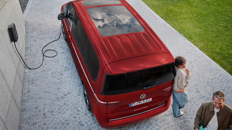 Vue aérienne d’un VW Multivan rouge en charge devant une maison via une borne murale, avec deux adultes sur l’allée pavée.