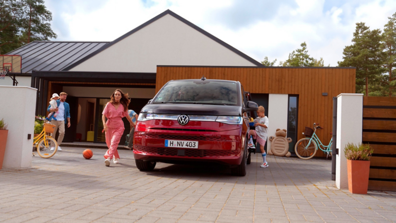 Vue de face d’un VW Multivan rouge devant une maison individuelle moderne ; la famille se déplace autour du véhicule pendant que les enfants jouent dans l’allée.