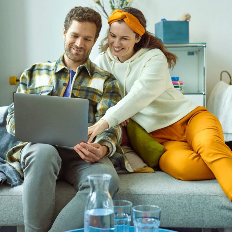 Ein Mann und eine Frau sitzen auf einer Couch und gucken einen Laptop Bildschirm an.