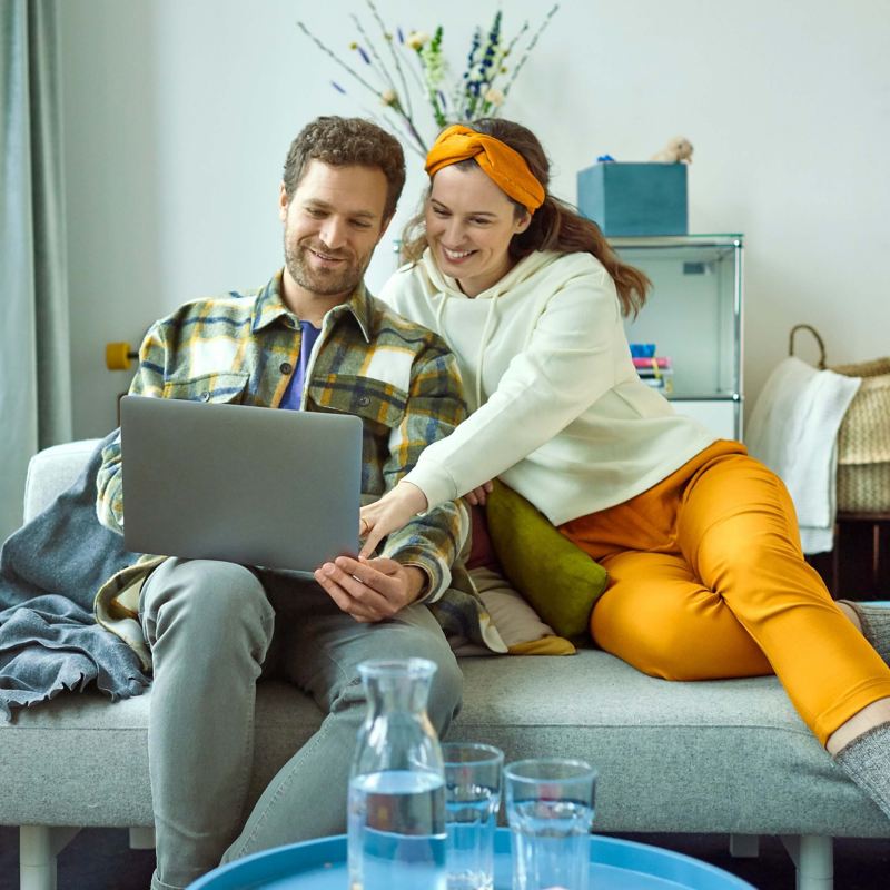 Ein Mann und eine Frau sitzen auf einer Couch und gucken einen Laptop Bildschirm an.