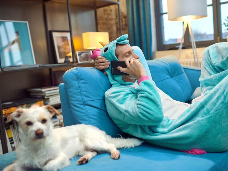 Ein Mann in einem Einhornkostüm sitz auf dem Sofa und telefoniert. Ein Hund liegt neben ihm.