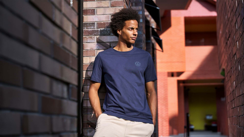 Hombre apoyado en un muro de ladrillo con playera azul marino y logo VW.